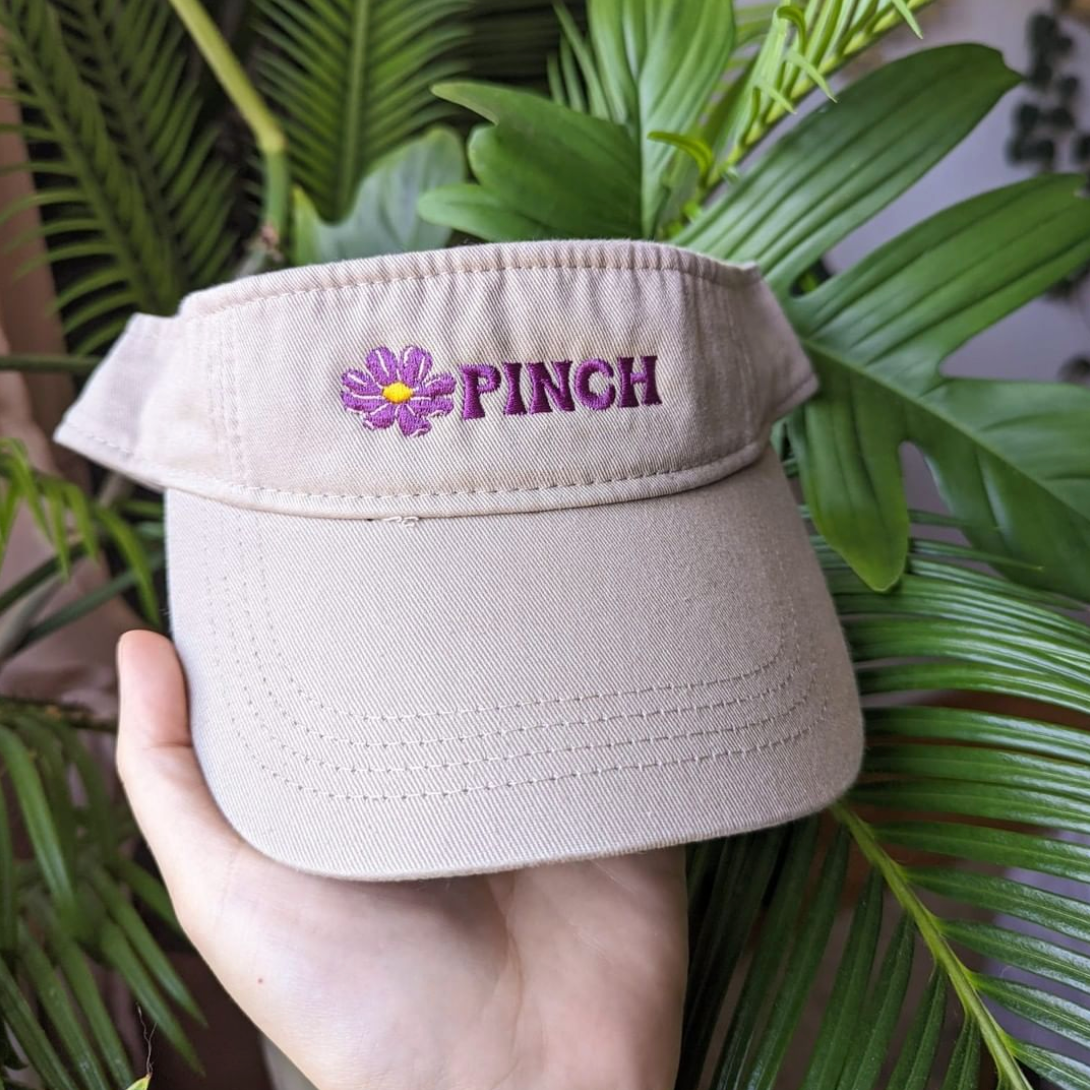 A light peach coloured hand holds a beige visor, embroidered with a dark pink flower and PINCH logo type. In the background is a large bright green fern.